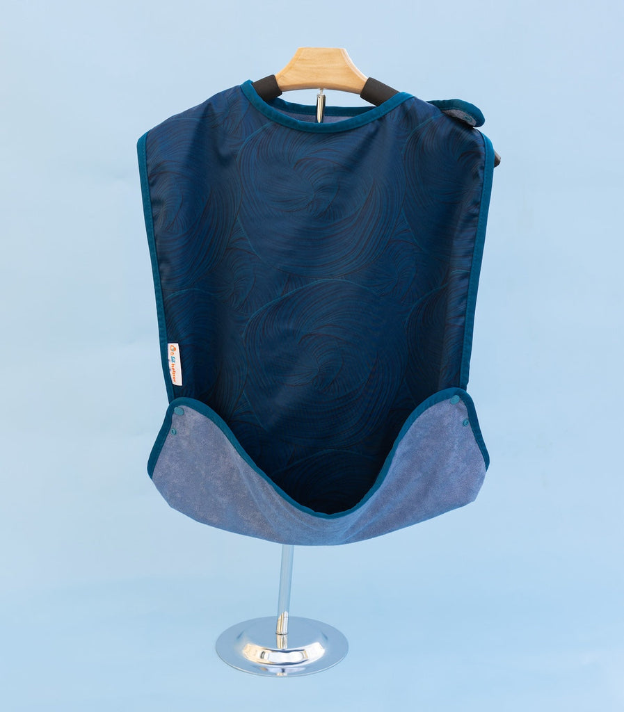 A Super Smock reusable adult bib rests on a clothes hanger. The bib is in a subtle dark blue pattern and features an optional pocket for catching crumbs.