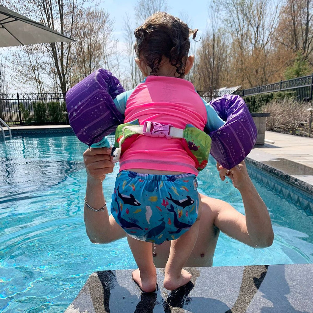 a toddler wearing a swim diaper and puddle jumper getting ready to jump into a pool with the help of an adult